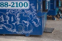 A blue dumpster features graffiti with various white, abstract designs and scribblings. The dumpster, with a partially visible telephone number, is situated on a concrete or gravel ground. The foreground shows some scattered debris and small objects.