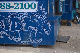 A blue dumpster features graffiti with various white, abstract designs and scribblings. The dumpster, with a partially visible telephone number, is situated on a concrete or gravel ground. The foreground shows some scattered debris and small objects.