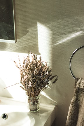 A bunch of dried lavender flowers are placed in a transparent glass jar on a white ceramic sink. The scene is illuminated by natural light streaming in from the left, casting soft shadows on the textured beige wall. A towel hangs on a circular metal rack visible on the right side.