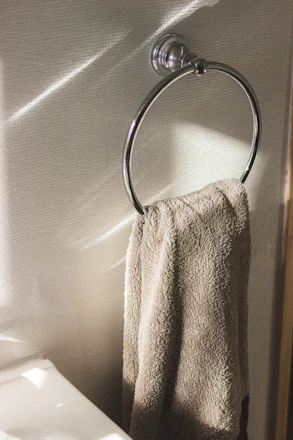 A chrome towel ring is mounted on a wall, holding a beige towel. Light from a window casts patterns on the wall and towel, creating a calm and peaceful atmosphere.