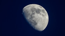 A detailed close-up view of the moon displaying its surface features, including craters and varying shades of gray. The moon appears on a clear, deep navy blue background, highlighting its bright white and gray tones.