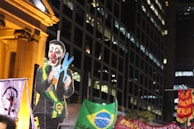 A nighttime protest scene features a large cutout of a figure with a clown face, wearing a suit and a ceremonial sash, holding oversized scissors. The background includes tall buildings and several banners, one displaying the Brazilian flag.