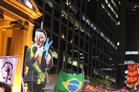 A nighttime protest scene features a large cutout of a figure with a clown face, wearing a suit and a ceremonial sash, holding oversized scissors. The background includes tall buildings and several banners, one displaying the Brazilian flag.