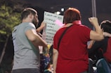 A group of people gathered around a person holding a sign with multiple options, such as education and feminism, written on it. The focus is on the interaction and communication among the individuals.