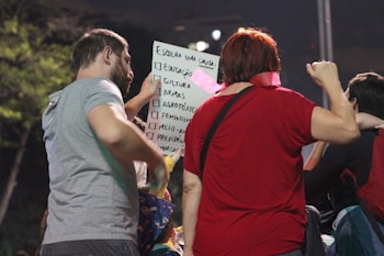 A group of people gathered around a person holding a sign with multiple options, such as education and feminism, written on it. The focus is on the interaction and communication among the individuals.
