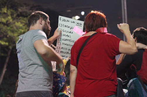 A group of people gathered around a person holding a sign with multiple options, such as education and feminism, written on it. The focus is on the interaction and communication among the individuals.