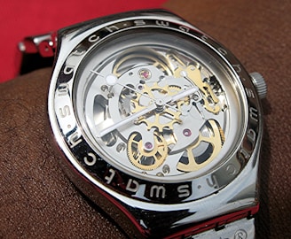 A close-up view of a mechanical wristwatch with visible gears and intricate components. The metal casing is shiny with the brand name inscribed around the bezel. The watch hands are thin and precise, positioned over the exposed internal workings of the watch.