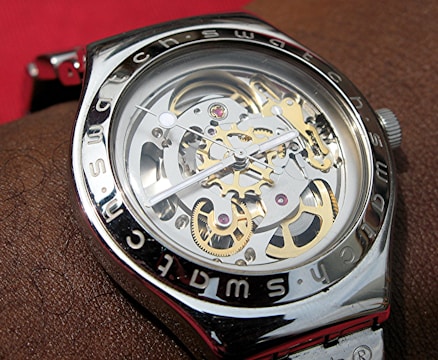 A close-up view of a mechanical wristwatch with visible gears and intricate components. The metal casing is shiny with the brand name inscribed around the bezel. The watch hands are thin and precise, positioned over the exposed internal workings of the watch.