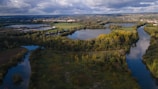 An aerial view of a lush landscape featuring rivers and lakes surrounded by dense greenery. Fields and clusters of trees are visible, with patches of sunlight breaking through the clouds, which cover much of the sky above. In the distance, there is a small town or village alongside farmlands and rolling hills.