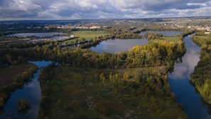 An aerial view of a lush landscape featuring rivers and lakes surrounded by dense greenery. Fields and clusters of trees are visible, with patches of sunlight breaking through the clouds, which cover much of the sky above. In the distance, there is a small town or village alongside farmlands and rolling hills.