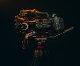A professional video camera mounted on a tripod is displayed against a dark background. The camera is equipped with various accessories, including handles and a lens, indicating it's suitable for high-quality video production.