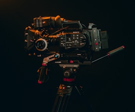 A professional video camera mounted on a tripod is displayed against a dark background. The camera is equipped with various accessories, including handles and a lens, indicating it's suitable for high-quality video production.