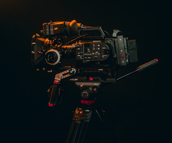 A professional video camera mounted on a tripod is displayed against a dark background. The camera is equipped with various accessories, including handles and a lens, indicating it's suitable for high-quality video production.