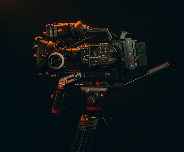 A professional video camera mounted on a tripod is displayed against a dark background. The camera is equipped with various accessories, including handles and a lens, indicating it's suitable for high-quality video production.