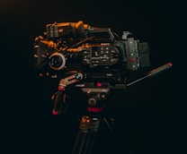 A professional video camera mounted on a tripod is displayed against a dark background. The camera is equipped with various accessories, including handles and a lens, indicating it's suitable for high-quality video production.