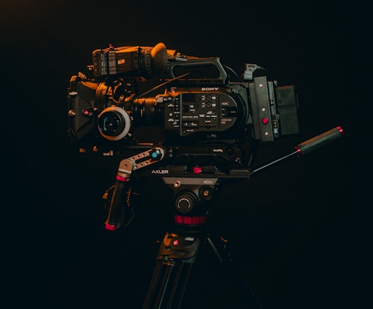 A professional video camera mounted on a tripod is displayed against a dark background. The camera is equipped with various accessories, including handles and a lens, indicating it's suitable for high-quality video production.
