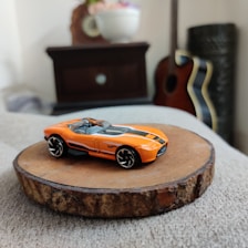 A bright orange toy sports car with black and white stripes is placed on a round wooden slab. In the background, there are household items such as a flower vase on a dark piece of furniture, a guitar with a black and white trim, and another object that appears to be a decorative column.