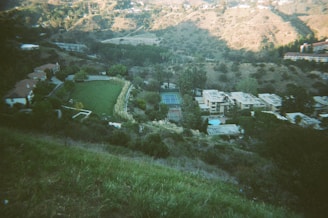 A scenic view from a hillside overlooking a residential area with several buildings and houses. The landscape includes lush greenery, a tennis court, and a lawn, surrounded by mountainous terrain. Trees and vegetation are abundant, with sunlight casting shadows on the landscape.