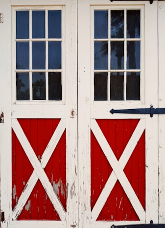 Old wooden double doors with a rustic, weathered look featuring red panels and white X-cross patterns. The doors include small, rectangular glass panes at the top, offering a view of tall trees outside.