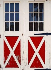 Old wooden double doors with a rustic, weathered look featuring red panels and white X-cross patterns. The doors include small, rectangular glass panes at the top, offering a view of tall trees outside.