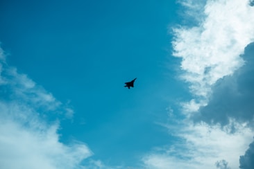 A silhouette of a jet fighter soaring through a bright, partly cloudy blue sky. Fluffy white clouds frame the scene with the jet prominently positioned in the center.
