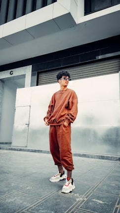 A person wearing a matching brown tracksuit and white sneakers is standing casually in an urban setting. The person has curly hair and is wearing sunglasses. The background includes a metallic wall and a partially closed roller shutter, with a modern building above.
