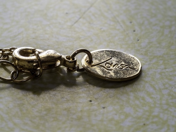 A close-up view of a gold-colored metallic necklace clasp attached to an oval pendant. The pendant has engraved lettering on its surface. The background is a muted, textured surface that contrasts with the shiny metal.