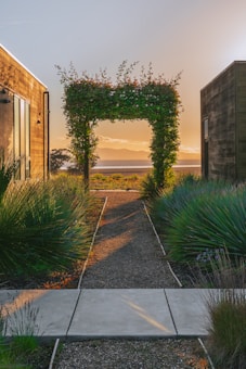 A pathway leads between two modern wooden buildings toward an archway covered in greenery. Beyond the archway, there is a scenic view of a body of water and mountains in the background, bathed in the warm glow of the setting sun. The surrounding plants and shrubs are lush and well-maintained.