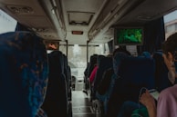 The interior of a bus where passengers are seated. A television screen at the front shows a scene from a movie or documentary, with a person in a green environment. Blue patterned upholstery covers the seats. A few passengers are visible, with one using a smartphone. The atmosphere appears calm and the lighting is subdued.