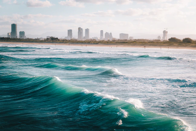 A coastal scene featuring striking turquoise ocean waves rolling towards a sandy shore. In the background, a line of modern high-rise buildings runs along the coastline, set against a partly cloudy sky. The beach appears to have small figures walking along it, indicating a leisurely atmosphere.