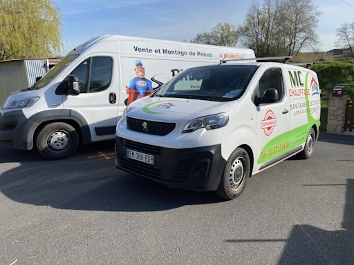 Two white vans are parked outdoors on a paved surface under a clear sky. The van on the left has branding related to tire sales and service in French. The van on the right displays advertisements for heating, sanitation, electrical, and central vacuum services. Both vans have signage and contact information on their sides.