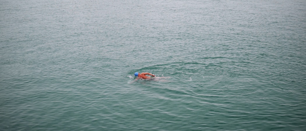 A single swimmer wearing a blue swim cap and goggles is moving through calm, open water. The swimmer is using a freestyle stroke and the water appears calm, expansive, and blue-green in color.