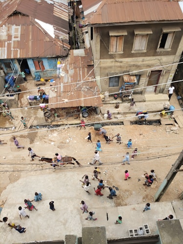 A street scene with numerous children walking in groups along a dirt pathway in front of old, weathered buildings with corrugated metal roofs. The buildings have a rustic appearance, with some wooden shutters and various household items visible. A horse with a rider can be seen among the children. Electrical wires hang overhead, and some people appear to be dressed in colorful clothing.