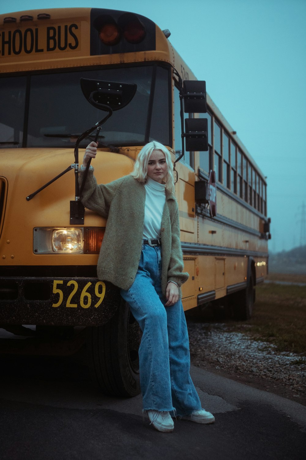 woman in gray sweater and blue denim jeans standing beside yellow bus during daytime