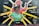 A vibrant mural features two figures with bold expressions, highlighting themes of justice and community empowerment. The foreground figure is wearing a green shirt with pins that read 'Defend Freddie Gray' and 'Save Mumia'. Behind, a figure in a pink suit raises a fist. Radiant yellow beams surround the figures, and words like 'Respect', 'Freedom', 'Peace', and 'Justice' are scattered across the background. The mural includes names and phrases referencing social justice and historical figures.