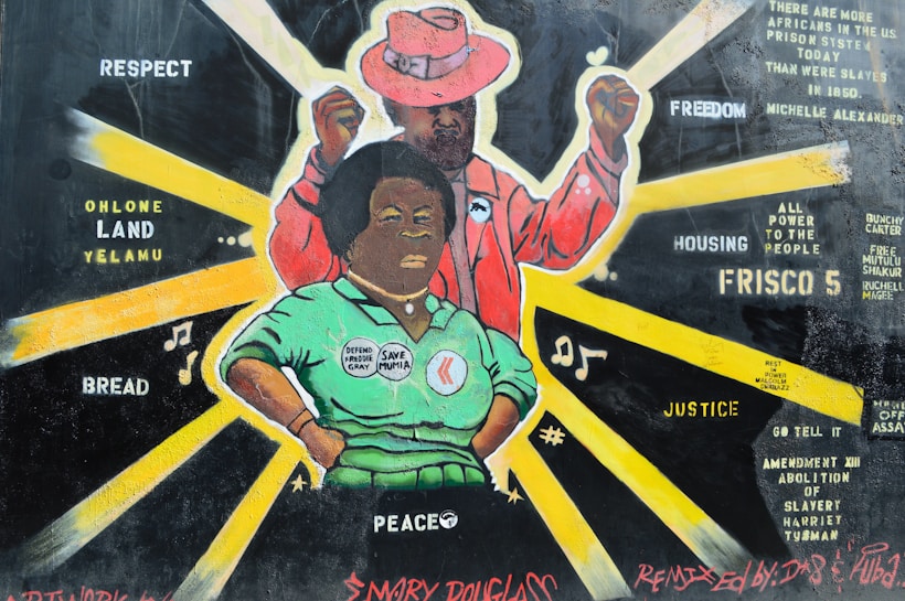 A vibrant mural features two figures with bold expressions, highlighting themes of justice and community empowerment. The foreground figure is wearing a green shirt with pins that read 'Defend Freddie Gray' and 'Save Mumia'. Behind, a figure in a pink suit raises a fist. Radiant yellow beams surround the figures, and words like 'Respect', 'Freedom', 'Peace', and 'Justice' are scattered across the background. The mural includes names and phrases referencing social justice and historical figures.