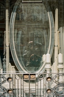 A large oval mirror reflects a street scene with an ornate building in the background. The mirror's frame is detailed with intricate designs and is supported by decorative columns. A faint, ghostly reflection of a person is visible in the glass.