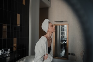 A person with a towel wrapped around their head and a bathrobe is standing in a bathroom. The background features a wall with dark tiles and a mirror. The person appears relaxed and is positioned near a bathtub and faucet.