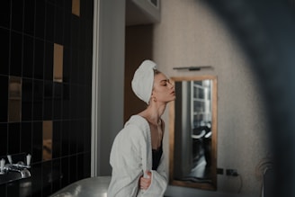 A person with a towel wrapped around their head and a bathrobe is standing in a bathroom. The background features a wall with dark tiles and a mirror. The person appears relaxed and is positioned near a bathtub and faucet.
