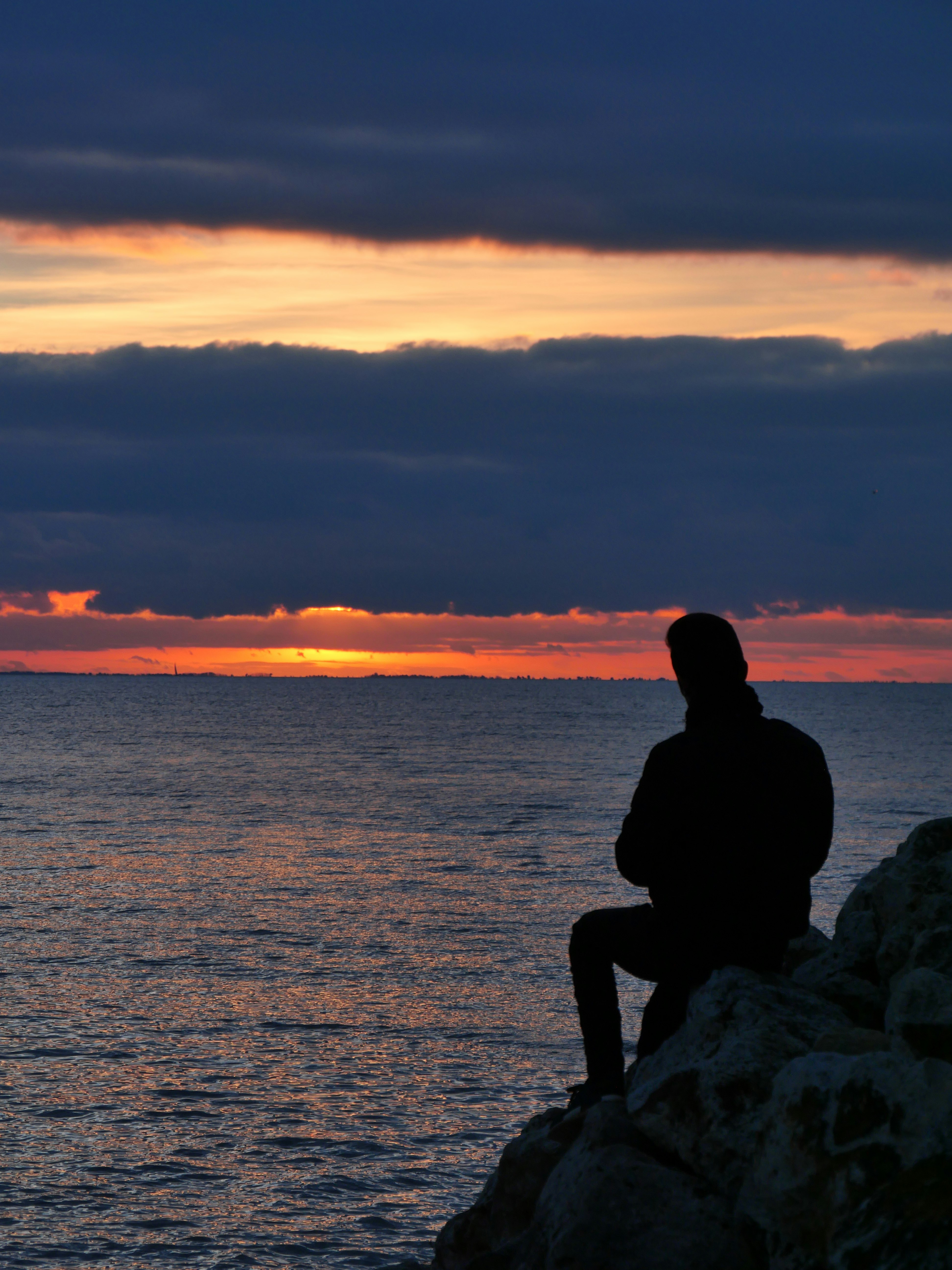silhouette of man sitting on rock near body of water during sunset