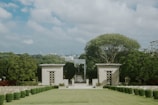 A landscaped area featuring well-maintained lawns with rows of shrubs and hedges, flanked by two square structures with geometric patterns. A large tree stands prominently behind the structures, and the cityscape can be seen in the background under a partly cloudy sky.