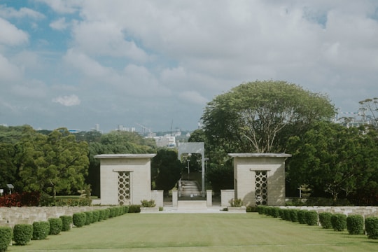 A landscaped area featuring well-maintained lawns with rows of shrubs and hedges, flanked by two square structures with geometric patterns. A large tree stands prominently behind the structures, and the cityscape can be seen in the background under a partly cloudy sky.