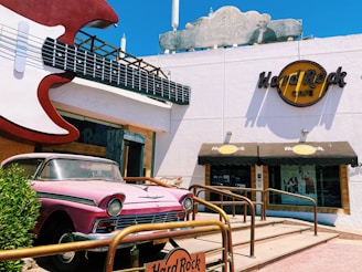 A building with a large guitar sculpture mounted at the entrance. A classic pink car is parked in front, next to a sign for the Hard Rock Cafe, which has a prominent logo on its facade. The building features a minimalist design with a beige and white color scheme, and there are railings leading to the entrance.