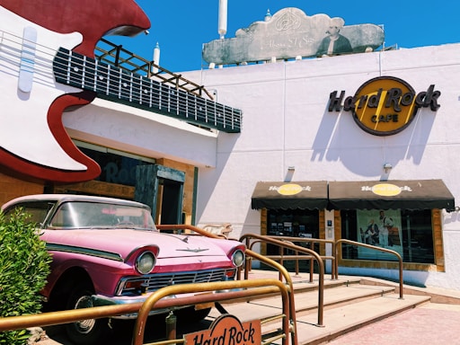 A building with a large guitar sculpture mounted at the entrance. A classic pink car is parked in front, next to a sign for the Hard Rock Cafe, which has a prominent logo on its facade. The building features a minimalist design with a beige and white color scheme, and there are railings leading to the entrance.