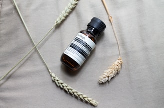 A small amber-colored bottle with a black cap lies on a textured beige surface, surrounded by wheat-like stalks. The bottle has a minimalist label with text, giving it an apothecary or skincare aesthetic.