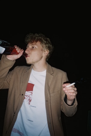 A person with short, curly hair is drinking from a bottle while holding a cigarette. They are wearing a tan blazer over a white T-shirt with red and black designs. The background is dark, implying nighttime or a dimly lit environment.