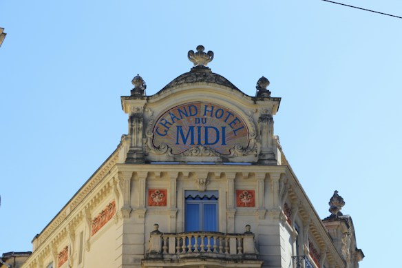 A decorative facade of an old building with architectural elements such as ornate carvings, a balcony with balustrades, and intricate detailing around the windows. At the top, there is signage with the text 'Grand Hotel du Midi' in blue letters on a white background. The building is set against a clear blue sky.