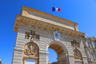 An ornate stone archway displays intricate carvings and sculptures, topped with the French flag against a clear blue sky. Latin inscriptions are engraved on the surface. The structure appears to be part of a historic or monumental building complex.