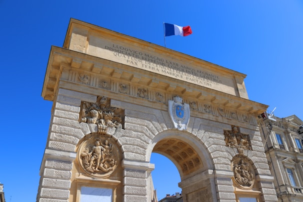 An ornate stone archway displays intricate carvings and sculptures, topped with the French flag against a clear blue sky. Latin inscriptions are engraved on the surface. The structure appears to be part of a historic or monumental building complex.