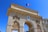An ornate stone archway displays intricate carvings and sculptures, topped with the French flag against a clear blue sky. Latin inscriptions are engraved on the surface. The structure appears to be part of a historic or monumental building complex.